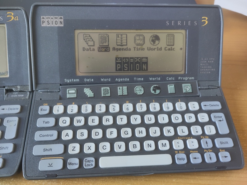 Psion series 3 palmtop computer. It looks like a mini laptop. The greyscale screen is showing a line of application icons.